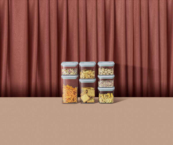 Set de recipiente transparente umplute cu alimente uscate, așezate pe o masă bej, în fața unui fundal cu perdea maro-roșiatică.