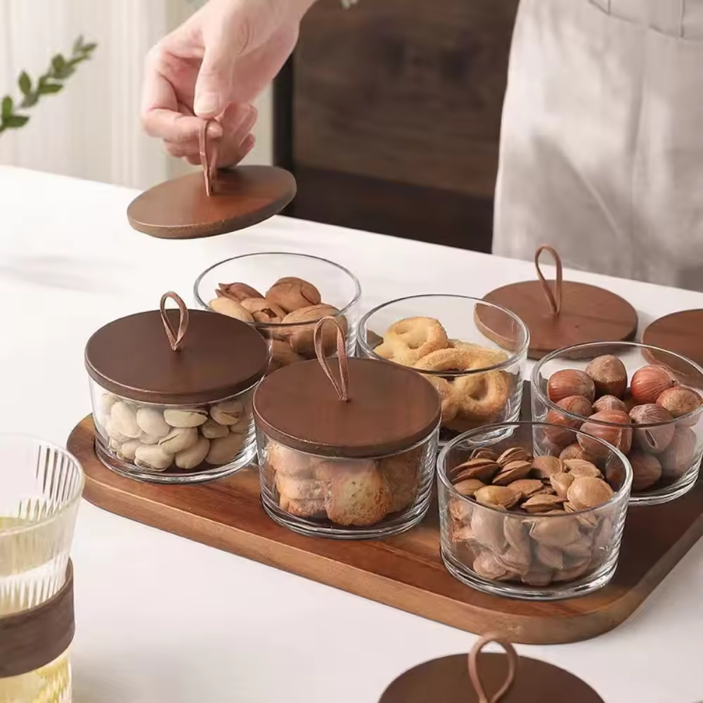 Persoană ridicând capacul unui borcan din sticlă cu capac de lemn, borcane pline cu gustări așezate pe tavă din lemn