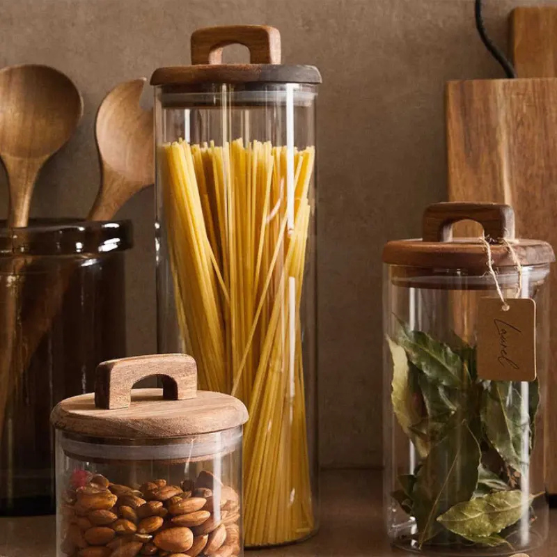 Set de 3 borcane din sticlă cu capac din lemn, umplute cu paste, migdale și frunze de dafin, perfecte pentru depozitarea alimentelor în bucătărie.