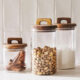 Set de 3 borcane din sticlă cu capac din lemn, umplute cu scorțișoară, fistic și făină, ideale pentru depozitarea condimentelor și alimentelor uscate în bucătărie.
