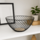Coș decorativ din metal negru cu bază din lemn, amplasat pe o etajeră albă modernă, lângă o ramă foto și o plantă decorativă.