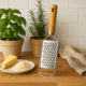 Răzătoare din inox cu mâner din lemn, așezată pe blatul din lemn lângă parmezan ras și plante aromatice