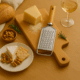 Răzătoare din inox cu mâner din lemn pe un platou de servire cu brânză, nuci, biscuiți și vin alb