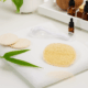 Burete exfoliant facial din lufă naturală, pe prosop alb, alături de accesorii cosmetice și frunze verzi